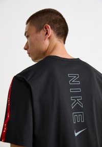 Melnā Nike T-kreklā ir apaļš kakls, īsas piedurknes un vertikāls pelēks logotips uz muguras. Piedurknes ir ar sarkaniem akcentiem un zīmola identifikāciju.