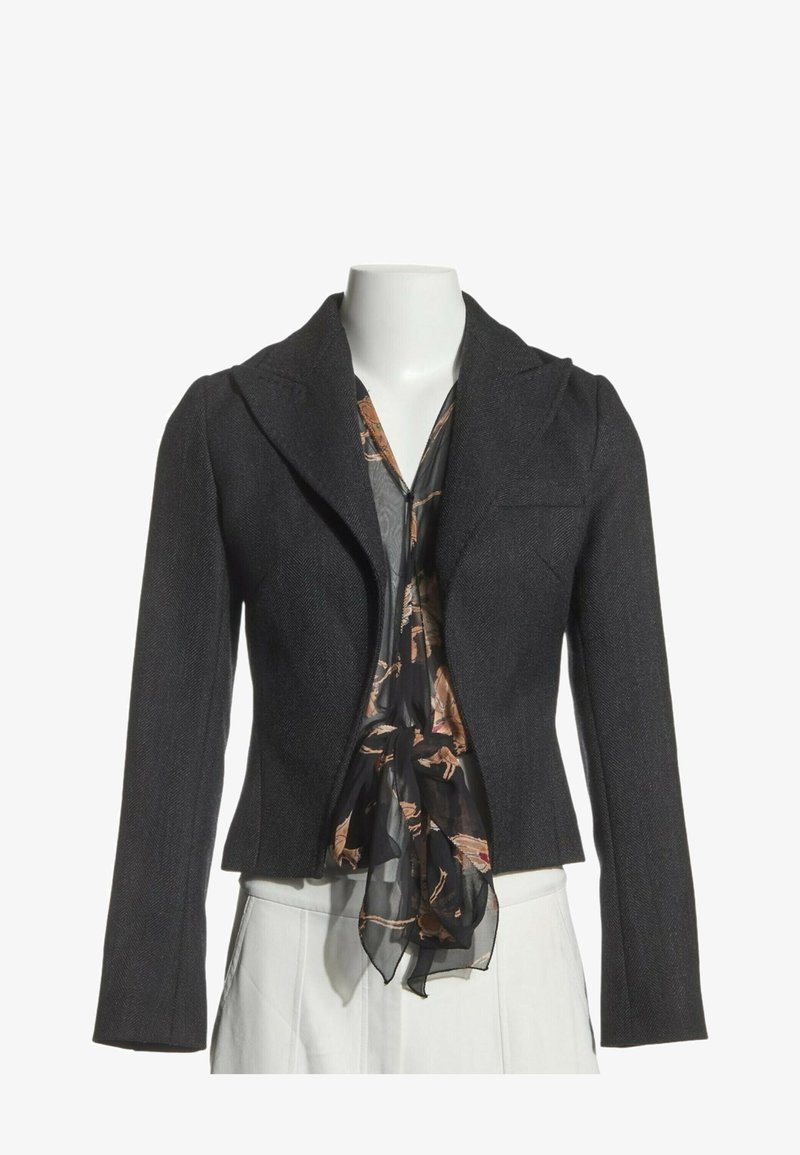 Veste cintrée gris foncé sur une écharpe noire à motifs et un pantalon blanc sur un mannequin sans tête sur fond blanc.