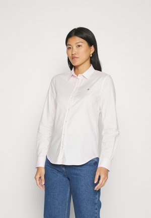 Camicia button-up rosa chiaro con maniche lunghe, colletto classico e un piccolo emblema sul lato sinistro del petto, abbinata a jeans blu.