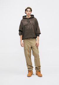 Felpa marrone con stampa grafica, pantaloni cargo beige e stivali color cuoio. L'outfit presenta una vestibilità comoda, tasca frontale e tasche laterali cargo.