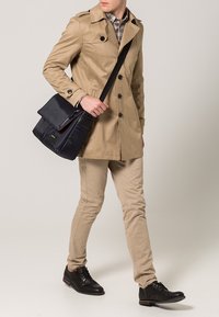 Trench coat beige con bottoni neri, indossato sopra una camicia a fantasia. Pantaloni beige chiaro e scarpe nere. Borsa a tracolla scura con dettagli in metallo.