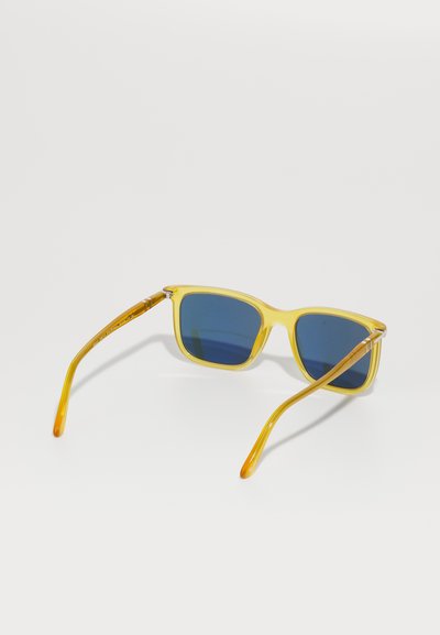 Óculos de sol de plástico amarelo com lentes azuladas. Design de armação quadrada, acentuado por hastes transparentes que transicionam para amarelo nas pontas.