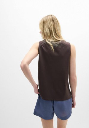 Blondýna otočená zády, oblečená v tmavě hnědém tílku bez rukávů a volných modrých šortkách, stojící před jednobarevným bílým pozadím.