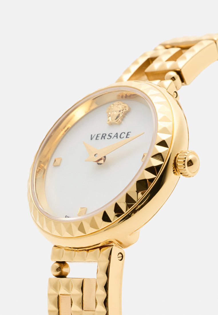 Montre dorée avec un boîtier rond texturé, un cadran blanc, un logo et des aiguilles dorées. Le bracelet à maillons présente un motif géométrique et une fermeture.