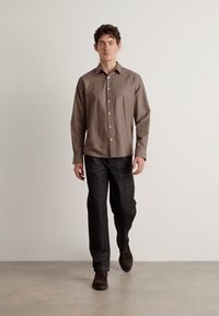 Cămașă maro cu nasturi și guler, asociată cu pantaloni din denim închis. Cămașa are o textură netedă, iar ținuta include pantofi maro.