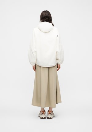 Personne portant une veste à capuche blanche, une longue jupe beige et des baskets beiges avec des accents orange, debout dos à nous sur un fond uni.