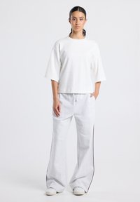 Woman standing against plain background wearing white oversized short-sleeve top and light gray wide-leg sweatpants with side stripes.