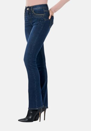 Donna che indossa jeans blu scuro decorati e stivaletti neri con tacco alto, in piedi di lato con una mano nella tasca posteriore.