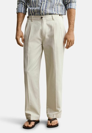 Man draagt beige geplooide broek, gestreept shirt met lange mouwen in de broek gestopt en donkere bruine teenslippers, staand tegen een effen achtergrond.