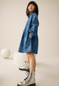 Denimkleid mit ausgestellt Rock und bestickten Schleifenmustern in Hellblau. Kombiniert mit glänzenden silbernen Schnürboots und einem runden weißen Objekt im Hintergrund.