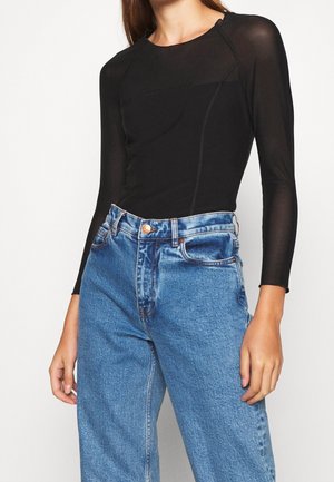 Vrouw die een aansluitende zwarte doorschijnende top met lange mouwen draagt, ingestopt in een high-waisted blauwe spijkerbroek, tegen een effen achtergrond.