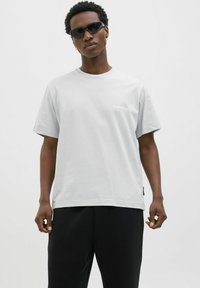 Lys grå kortærmet t-shirt i blødt bomuld med rund hals og lille hvid logo detalje på brystet; parret med sorte bukser.