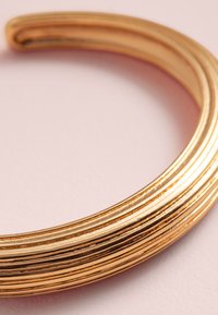 Goldfarbenes Metall-Manschettenarmband mit glatter Oberfläche und gerippter Textur, das eine abgerundete Form und einen dezenten Glanz vor einem rosa Hintergrund aufweist.