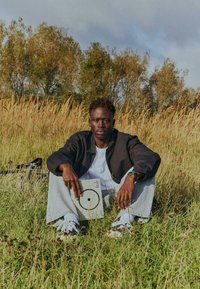 Personne assise dans une herbe haute, portant une veste noire et un jean clair, tenant un livre avec un motif circulaire, entourée par la nature.