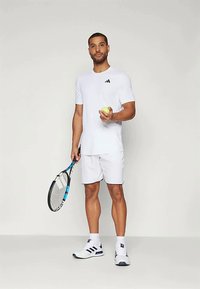 Camiseta deportiva blanca con logo negro, pantalones cortos blancos a juego, zapatillas de tenis blancas con acentos negros, sosteniendo una pelota de tenis amarilla y una raqueta.