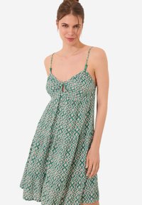 Robe verte à motif, avec une coupe évasée, des bretelles spaghetti, un détail de nœud à l'avant et un motif géométrique dans des couleurs contrastées.