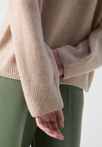 Personne portant un pull beige en tricot avec des poignets côtelés et un pantalon vert, une main pendant librement et l'autre partiellement visible.
