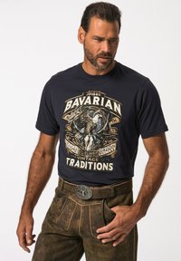 Námořnické tričko s potiskem, které zobrazuje centrální design orla a text, v kombinaci s hnědými texturovanými kalhotami s opaskem a ozdobnou sponou.