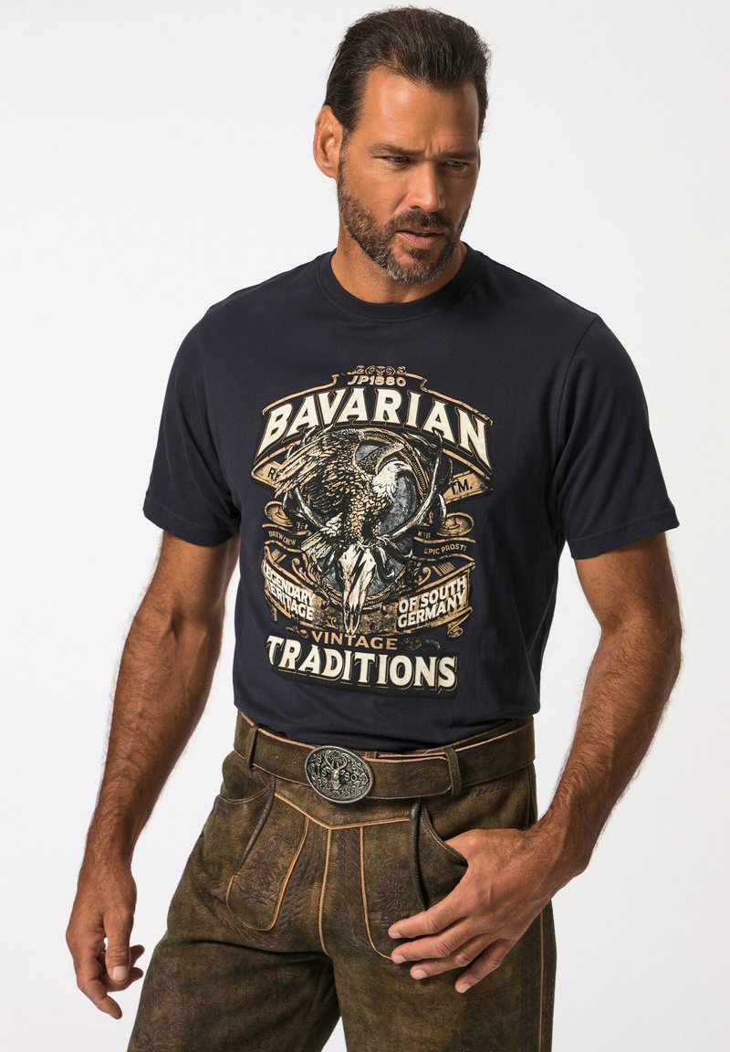 Námořnické tričko s potiskem, které zobrazuje centrální design orla a text, v kombinaci s hnědými texturovanými kalhotami s opaskem a ozdobnou sponou.