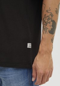 Sort tekstureret T-shirt med et rektangulært mærke i kanten, båret sammen med blå denim jeans; synlige arm-tatoveringer fremhæver looket.