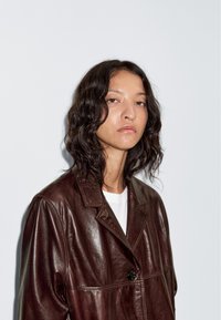 Grove bruine leren oversized jas met een gladde textuur, voorzien van een knoopsluiting en een reverskraag, gedragen over een wit T-shirt.