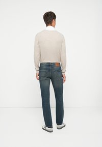 Suéter de canalé gris claro con mangas largas, combinado con jeans oscuros. Los jeans cuentan con un parche de cuero en la parte trasera. Se llevan sandalias.