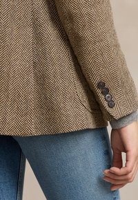 Brązowa marynarka w jodełkę z rękawem ozdobionym czterema czarnymi guzikami, noszona na szarej bluzie, w połączeniu z niebieskimi dżinsami, ręka swobodnie opadająca wzdłuż ciała.