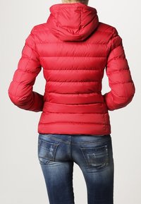 Rød pufferjakke med hætte, med horisontalt quiltede sektioner. Båret med tætsiddende blå jeans, der har en falmet vask og dekorativ syning.