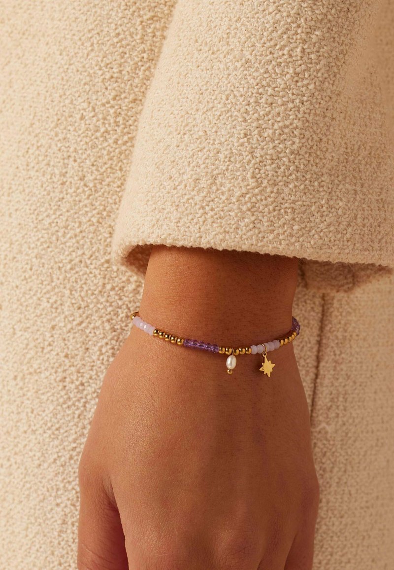 Delicate armband met gouden kralen, paarse en roze stenen, en een kleine gouden ster charme, gedragen op een pols met een textuur van stof als achtergrond.
