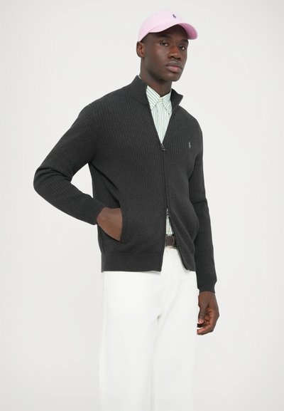 Sötétszürke bordázott cipzáras kardigán oldalzsebekkel, világoszöld csíkos ing fölött viselve, fehér nadrággal és rózsaszín sapkával kombinálva.