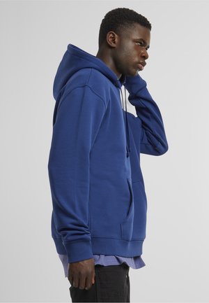 Jeune homme en sweat à capuche bleu et pantalon noir, debout de profil, une main touchant le côté de son cou, sur un fond uni.