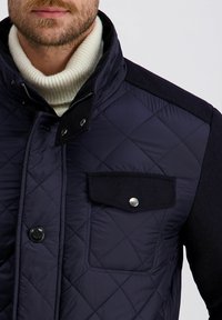 Mann in einer marineblauen Steppjacke mit einer geknöpften Brusttasche und einem weißen gerippten Rollkragenpullover.