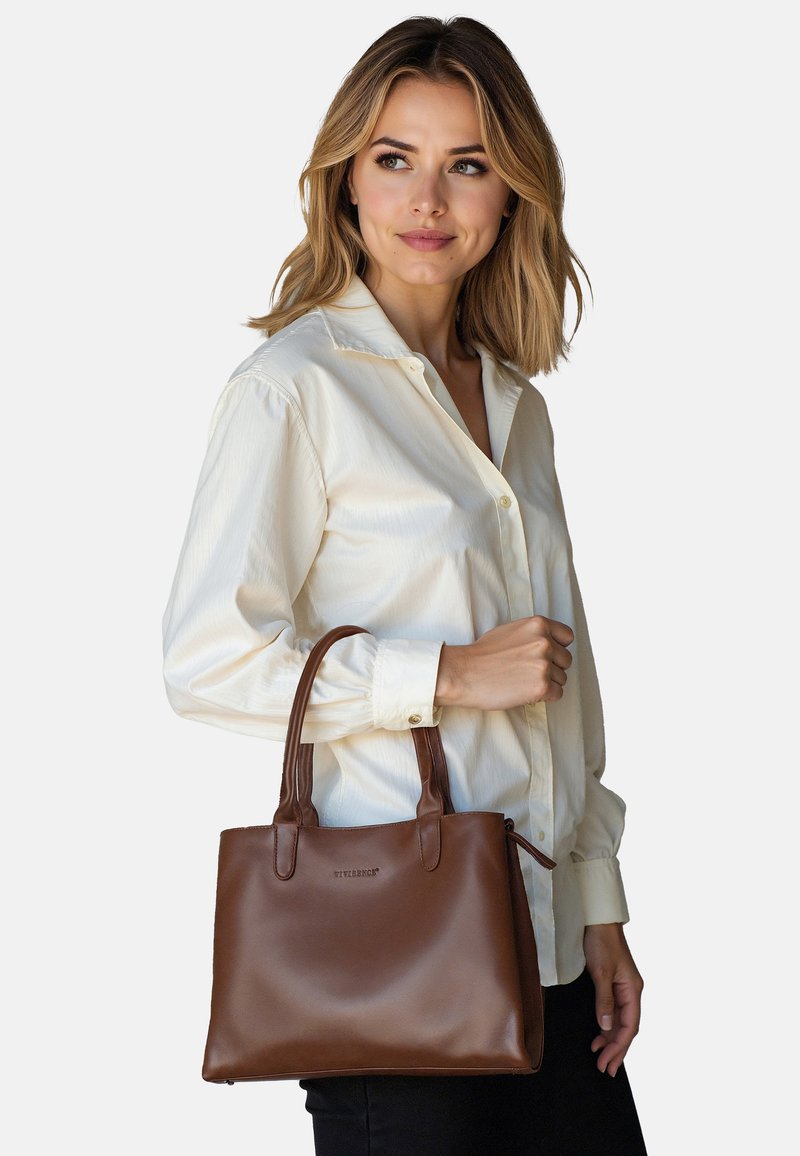 Mujer con blusa crema sostiene bolso de cuero marrón con asas dobles, mirando de lado contra un fondo liso.
