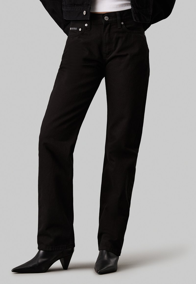 Vaqueros de mezclilla negra con corte recto, diseño de cinco bolsillos y herrajes de marca, combinados con tacones negros puntiagudos.