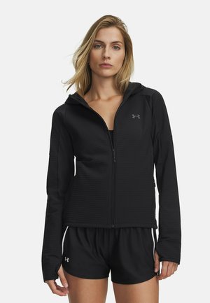 Vrouw draagt een zwarte hoodie met ritssluiting en zwarte sportshorts, staande tegen een effen witte achtergrond.