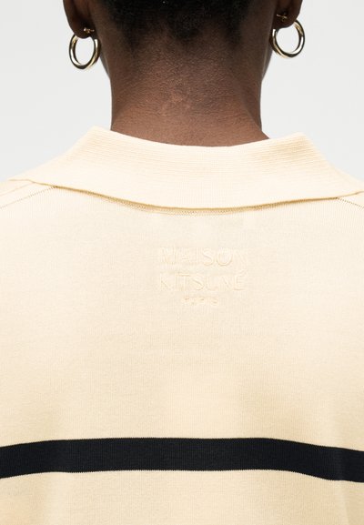 Πλάτη προσώπου που φοράει εκρού πλεκτό τοπ με μαύρη ρίγα και ανάγλυφη επιγραφή "Maison Kitsuné Paris", εμφανίζοντας χρυσούς κρίκους σκουλαρίκια.