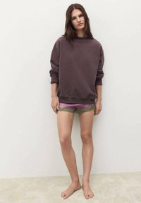 Sudadera marrón de gran tamaño, combinada con pantalones cortos morados claros que tienen un borde de encaje verde. Fondo sencillo, modelo descalzo.