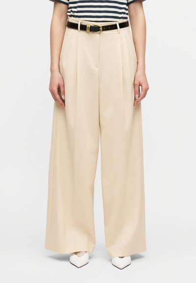 Femme portant un pantalon large beige avec des plis à l'avant, une ceinture noire avec une boucle dorée, et des chaussures blanches pointues avec des détails en dentelle.
