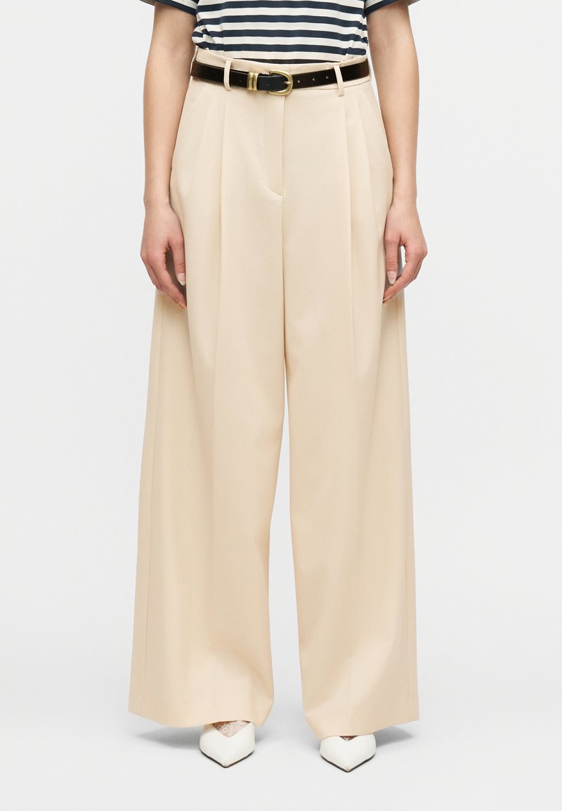 Γυναίκα που φοράει μπεζ παντελόνι  με φαρδιά πόδια και εμπρόσθιες πιέτες, μαύρη ζώνη με χρυσή αγκράφα και μυτερά λευκά παπούτσια με λεπτομέρειες δαντέλας.