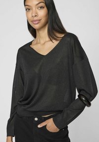 Musta, pitkähihoinen paita V-pääntiellä, joka on valmistettu pehmeästä, teksturoidusta kankaasta. Yhdessä hihassa on musta resori. Yhdistetty mustiin housuihin.