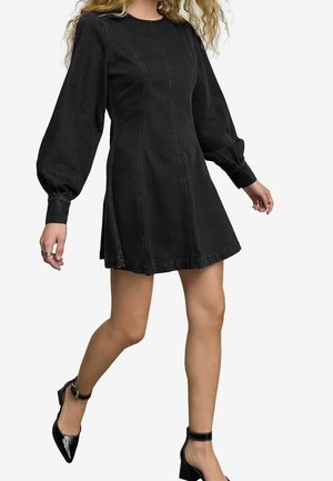 Czarna sukienka dżinsowa z długimi balonowymi rękawami, okrągłym dekoltem, pionowymi przeszyciami i rozkloszowanym dołem. Nosi ją z czarnymi szpilkami.