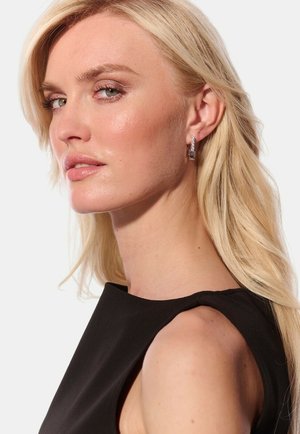 Sølv hoop øreringe med et rektangulært design, prydet med små klare perleaccent. Modellen har langt blondt hår og bærer en sort top.