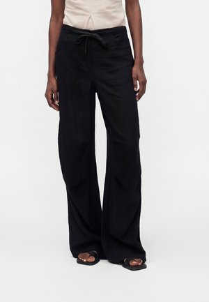 Personne portant un pantalon noir ample avec taille à cordon et des sandales noires, debout devant un fond blanc uni.