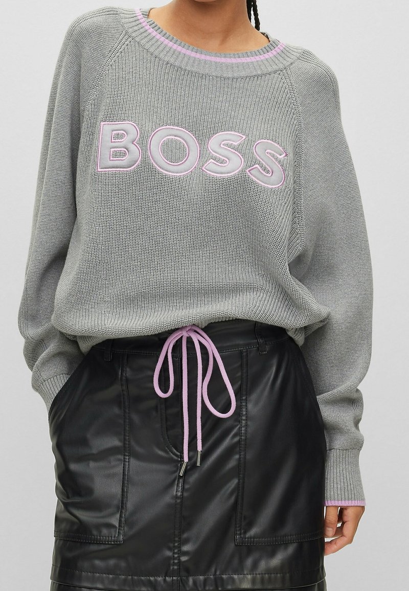 Grauer gestrickter Pullover mit pinker "BOSS"-Stickerei, ausgestattet mit einem gerippten Kragen und Bündchen, kombiniert mit einem schwarzen Lederrock mit Kordelzugakzenten.