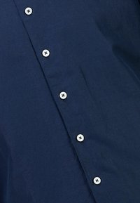 Donkerblauwe stof met een verticale rij van zes gelijkmatig verspreide witte knopen, genaaid met een donkere draad.