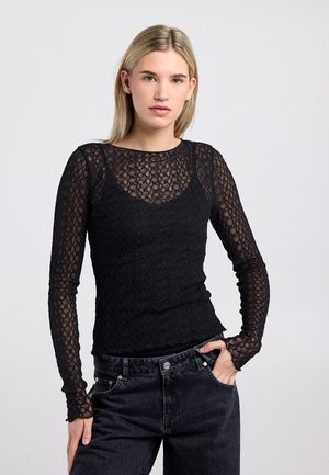 Haut noir à manches longues avec un motif en maille texturée, doté d'un décolleté arrondi et d'une silhouette ajustée. Associé à un jean foncé.