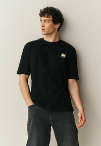 Kócos hajú férfi fekete Basquiat pólóban és sötét farmerben, balra néz, egyik kezét a zsebében tartja, bézs fal előtt.