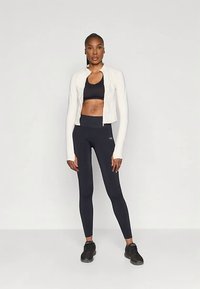 Nő áll, kezét az oldalán tartva, fekete sportmelltartót, magas derekú fekete leggingset, fehér rövid, zipzáras dzsekit és fekete sportcipőt viselve.