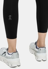 Černé sportovní legíny s viditelným logem a délku ke kotníkům, doplněné šedobílými běžeckými botami s výrazným odpružením a detaily šněrování.