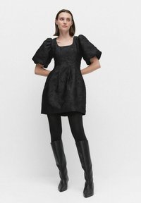 Svart puffärmad klänning med texturerat blommönster, fyrkantig halsringning, figursydd midja, kombinerad med svarta strumpbyxor och knähöga läderstövlar.